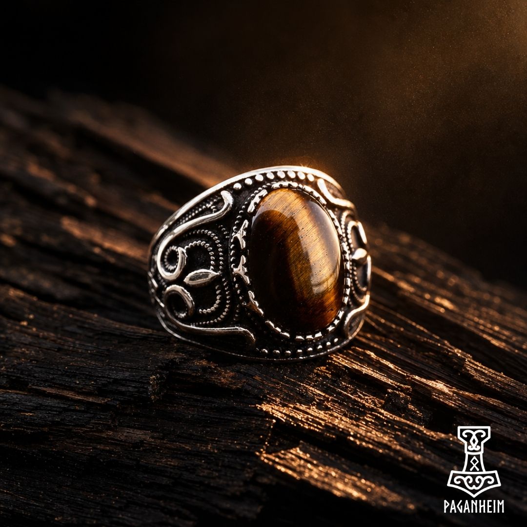 A silver-toned Viking-style ring with an oval brown tiger's eye stone, resting on a dark textured wooden surface with dramatic warm lighting.