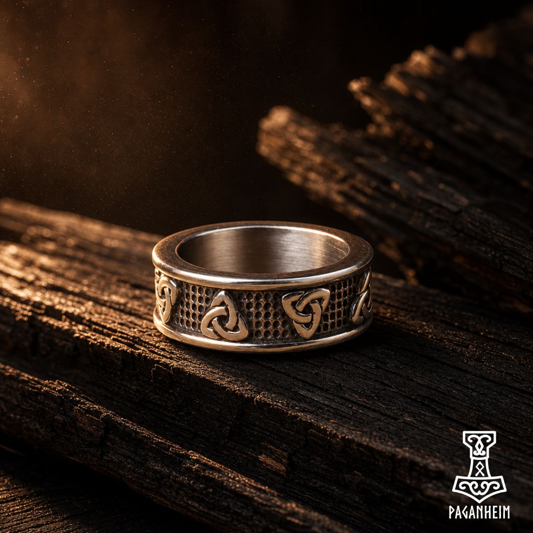 A silver-toned Viking-style ring with triple knot (triquetra) and wavy motifs, resting on dark weathered wood. The brand logo 'Paganheim' is visible in the bottom right corner.