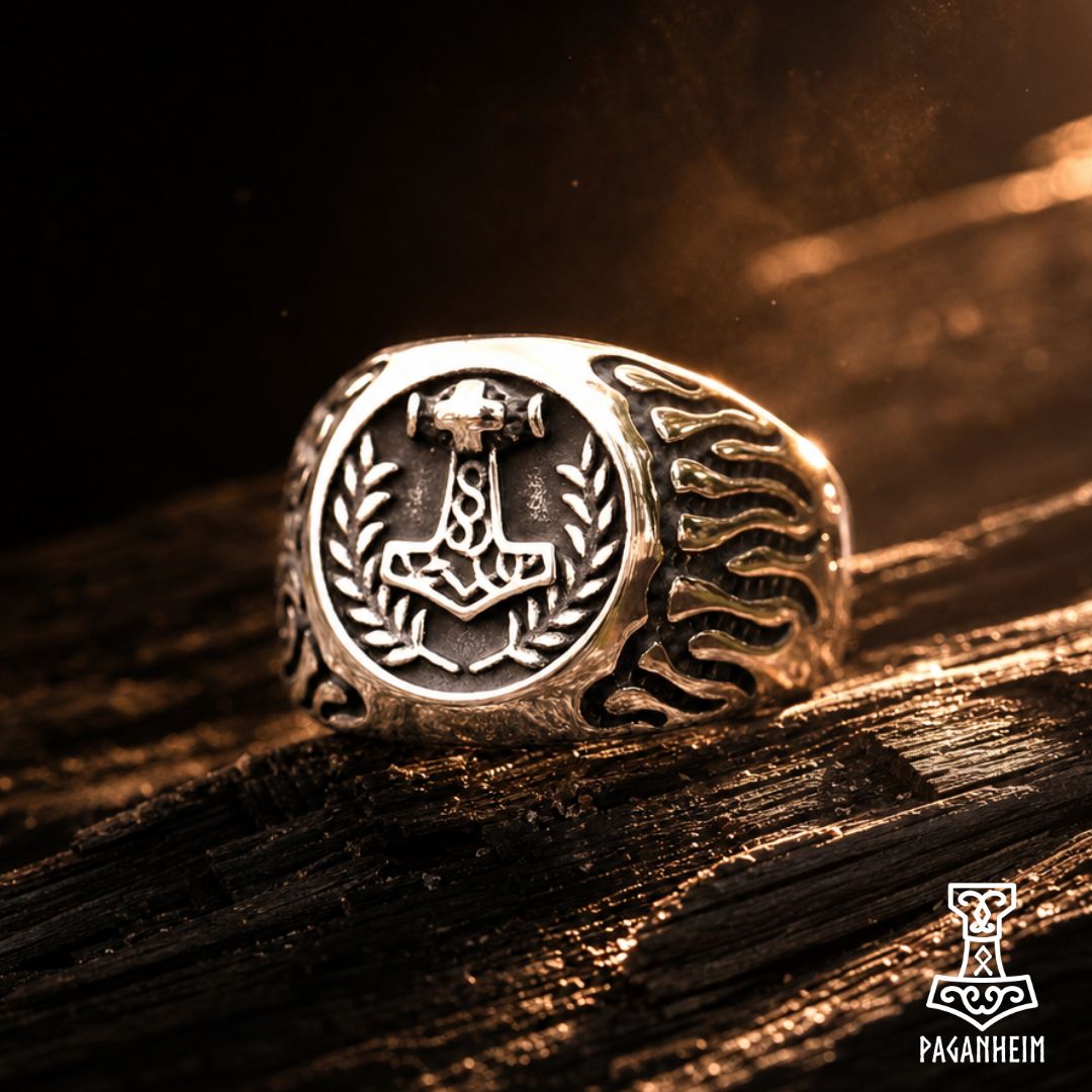 A silver-toned Norse ring featuring a hammer (Mjolnir) and laurel wreath emblem on the face, with a textured flame-like pattern on the band, resting on dark wood under dramatic golden lighting.