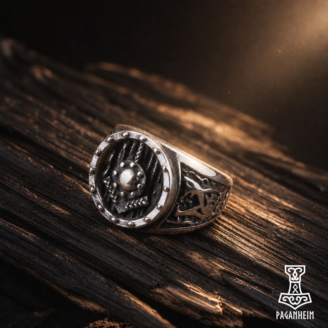 A silver-toned Viking-style ring featuring a skull motif on a circular face and a carved band, displayed on a dark textured wooden surface with dramatic warm lighting. The Paganhim logo is visible in the bottom right corner.