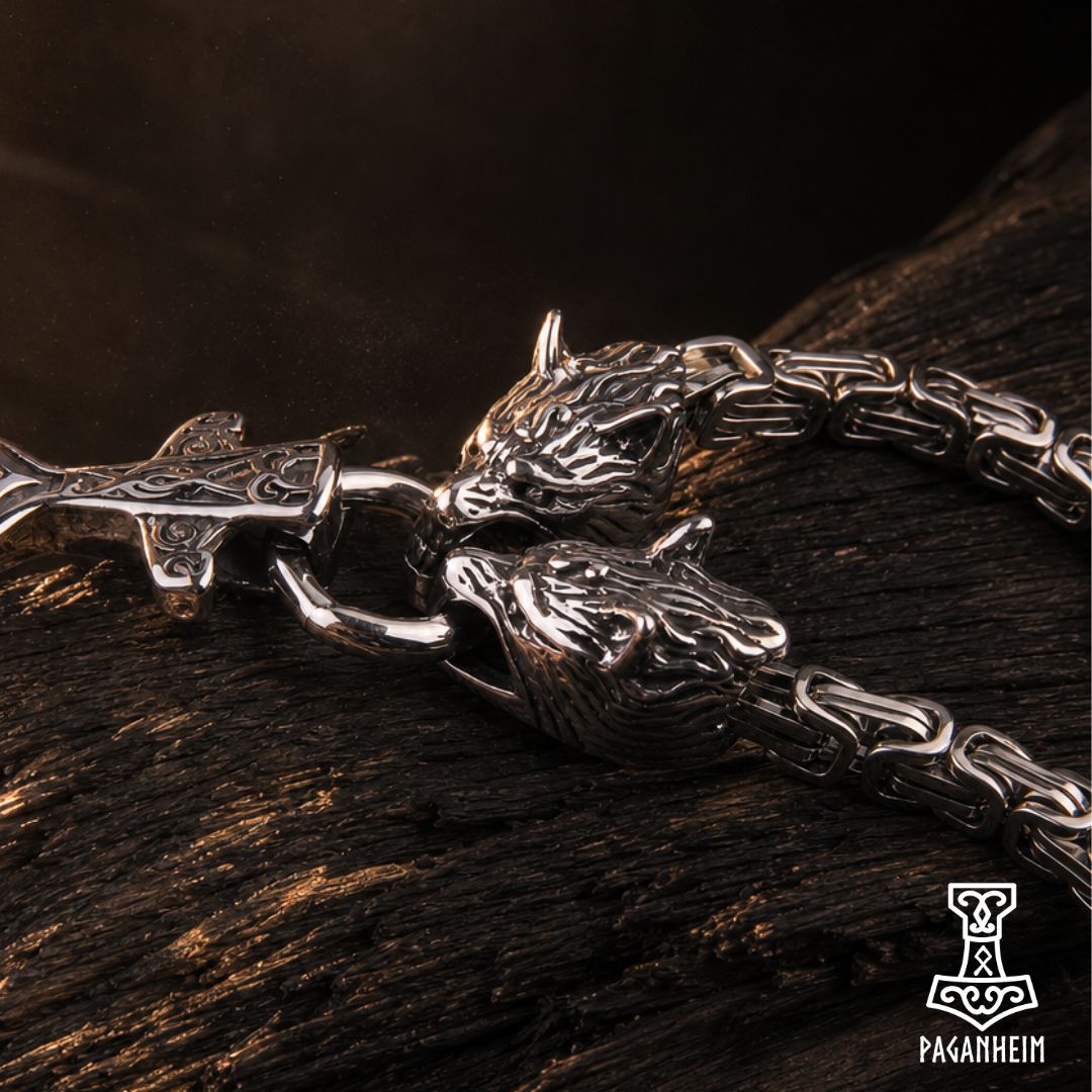 A close-up of a silver-toned Viking-style chain necklace with two detailed wolf head charms and an engraved pendant, resting on a dark textured wooden surface.